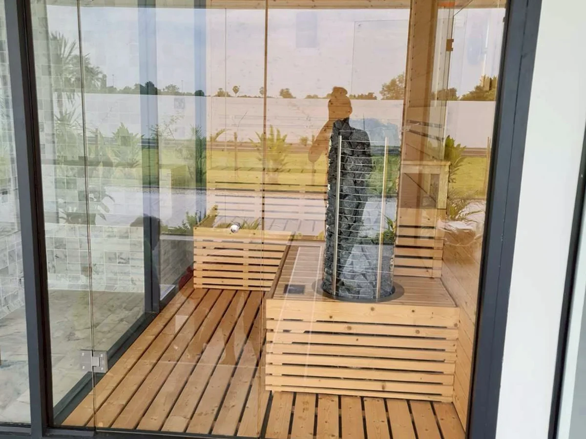 Glass-corner sauna with a stone-filled tower heater, garden and open green field beyond the glass