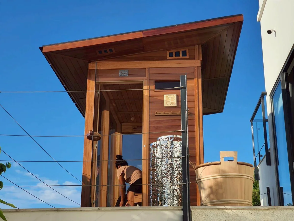 Rooftop cedar sauna tower with wide eaves against a blue sky, outdoor shower fitting beside the glass door