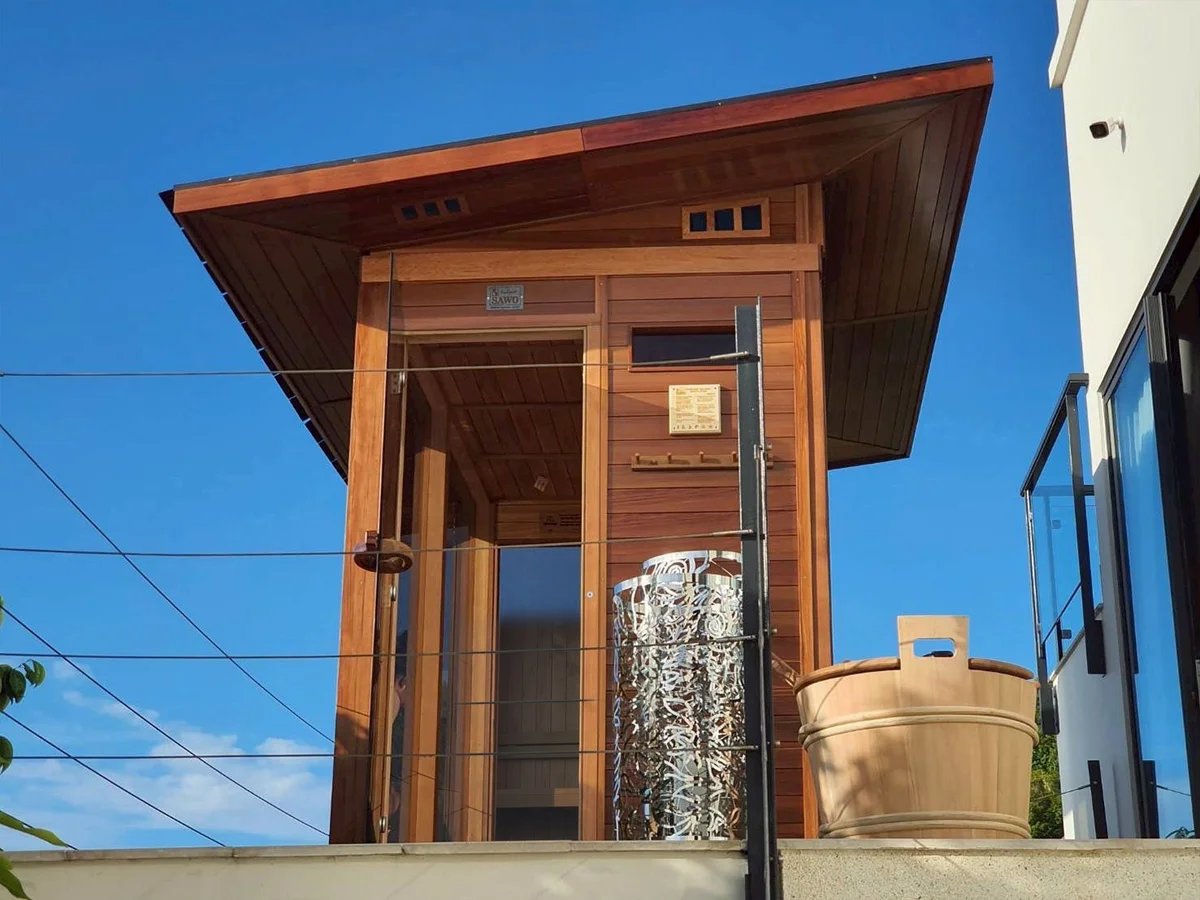 Rooftop cedar sauna tower with wide eaves against a blue sky, outdoor shower fitting beside the glass door