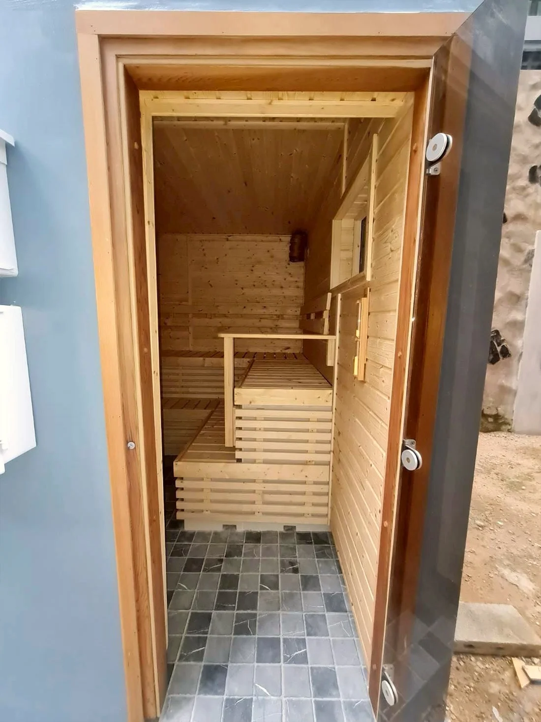 Small pale-timber sauna seen through its open door, slate floor in the foreground