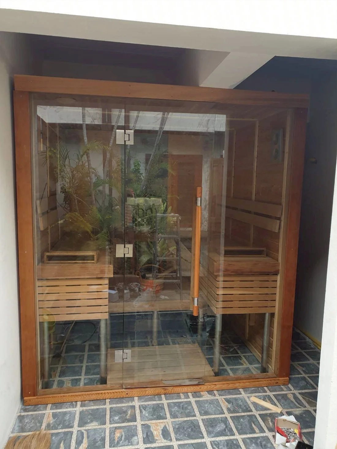 Teak-framed glass sauna standing in a tropical courtyard with greenery around it