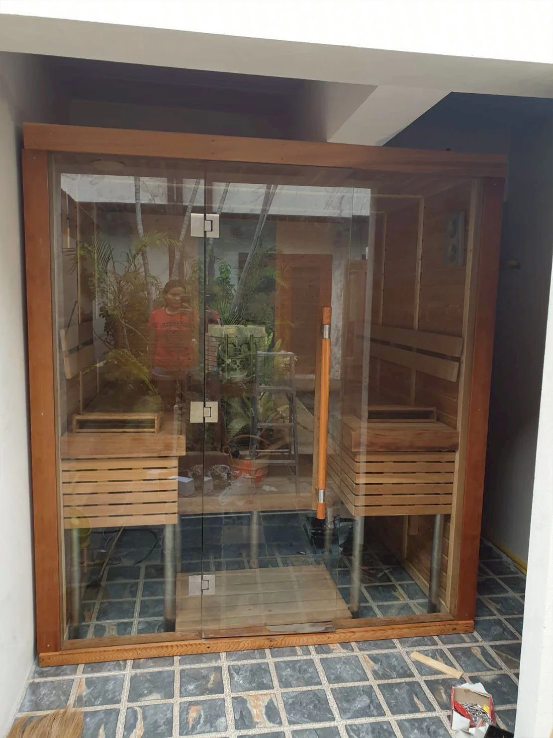 Teak-framed glass sauna standing in a tropical courtyard with greenery around it