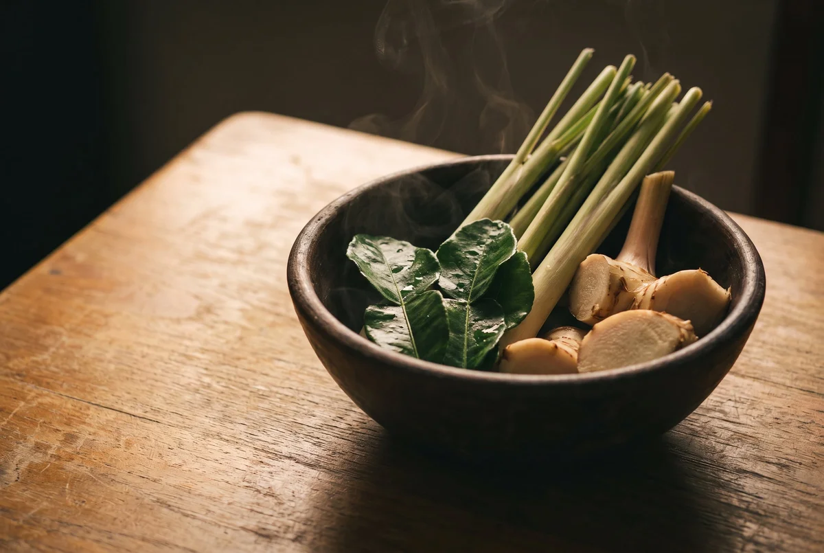 A bowl of Thai herbs — lemongrass, kaffir lime, galangal — with steam rising, the heritage bridge
